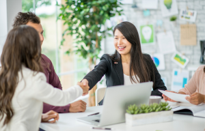 Two women shaking hands in a workplace setting with other team members around