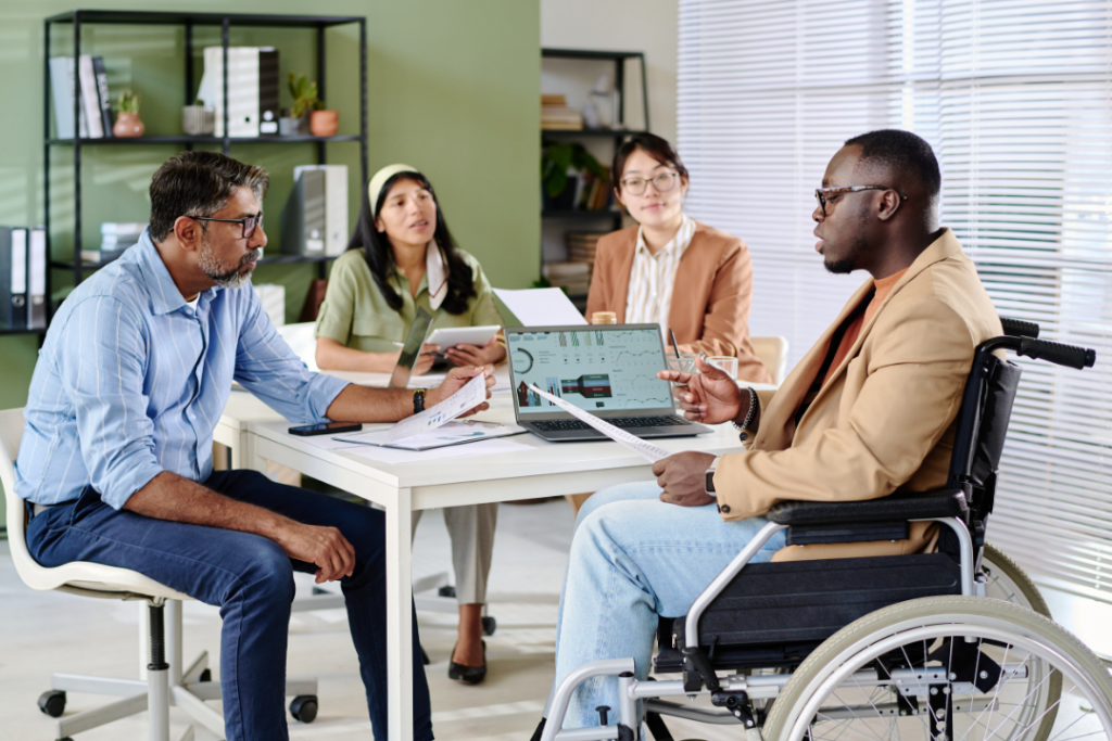 Group of business professionals sitting around table, engaging in important meeting. People reviewing documents, one person in wheelchair, modern office environment